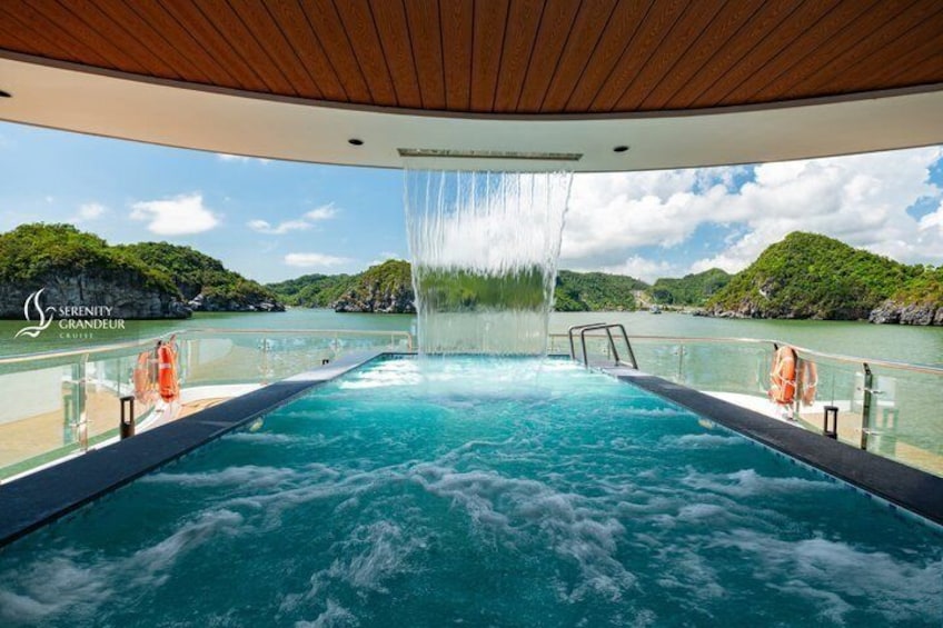 A pool with a waterfall feature, surrounded by a wooden deck and a railing, overlooking a body of water with greenery in the background. 2
