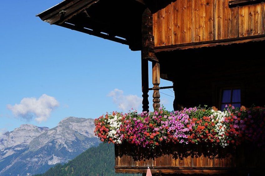 A wooden chalet with a balcony filled with colorful flowers, set against a backdrop of mountains. 1