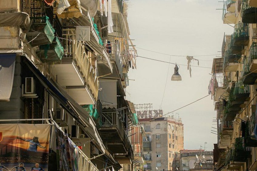A narrow urban street with multi-story buildings, balconies, and hanging laundry. 5