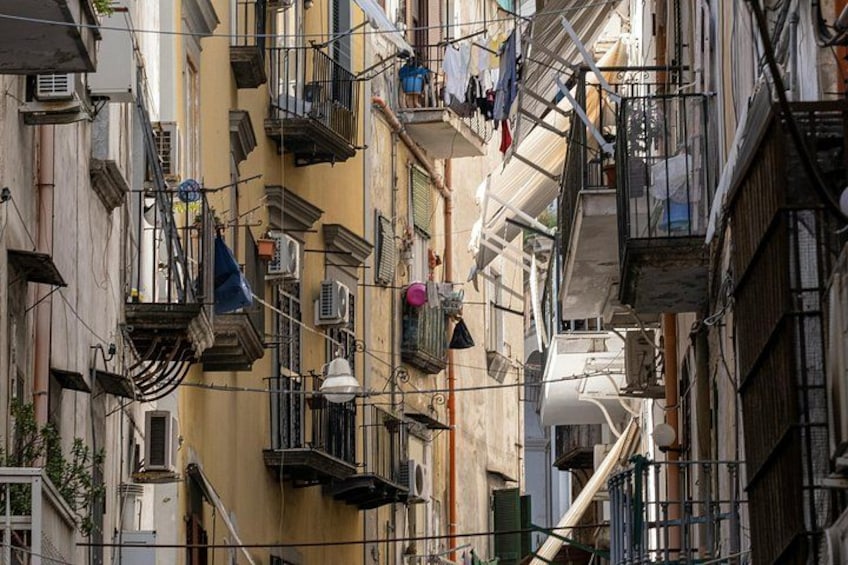 A narrow urban street with multi-story buildings, balconies, and hanging laundry. 4