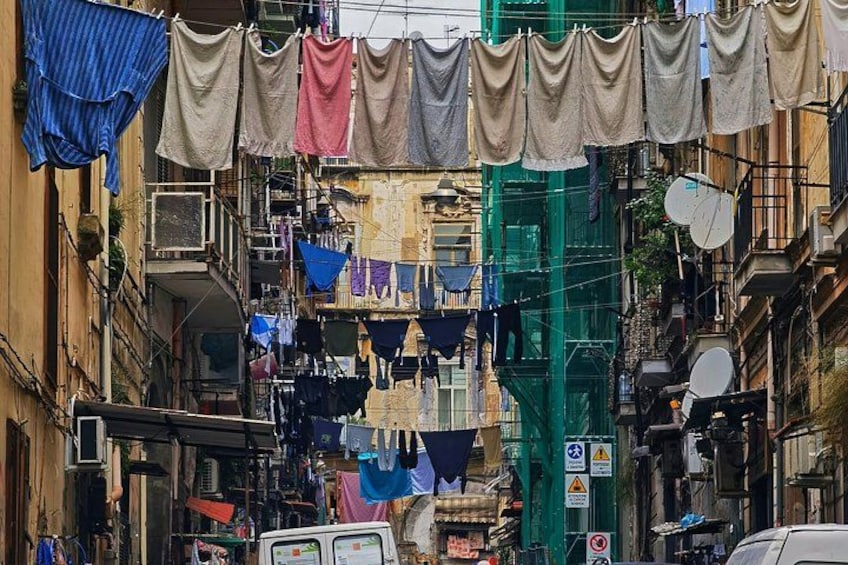 A narrow street with buildings on either side, laundry hanging between them, and a green construction crane. 2