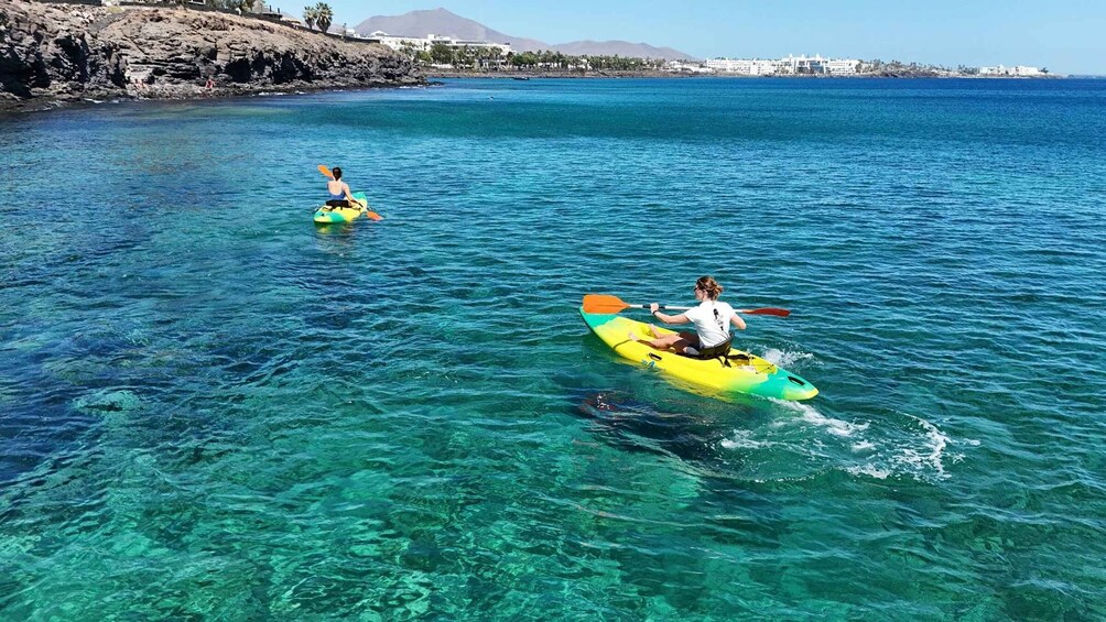 Two people kayaking on clear blue water with a rocky coastline and buildings in the background. 1