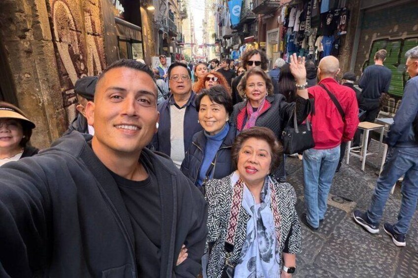 A crowded street with people walking and standing, historic stone buildings, and a narrow alleyway. 3