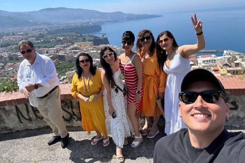 A group of people on a stone wall overlooking a coastal town with buildings and the sea in the background. 1