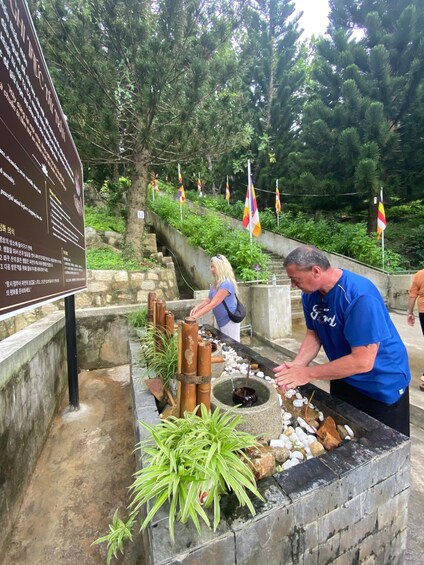 A man in a blue shirt operating a traditional water mill, with a signboard and flags in the background. 2