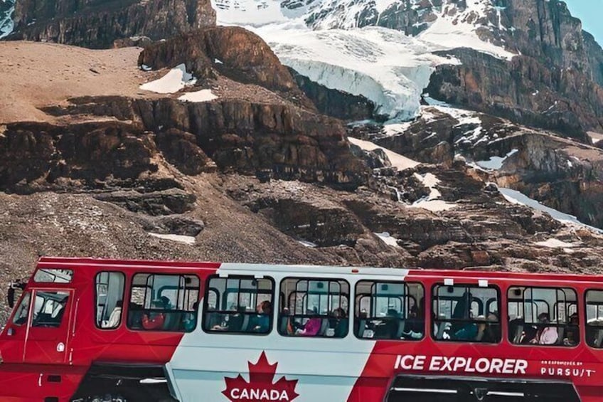 A red and white Canada Ice Explorer bus with passengers, traveling through a mountainous area with snow patches and a glacier in the background. 4