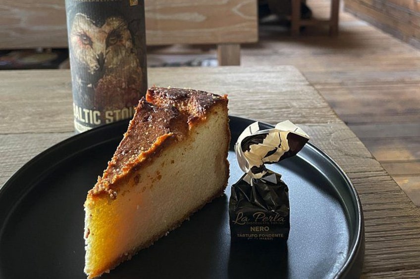 Una porción de pastel de queso sobre un plato negro, con una barra de chocolate y una botella al fondo. 3