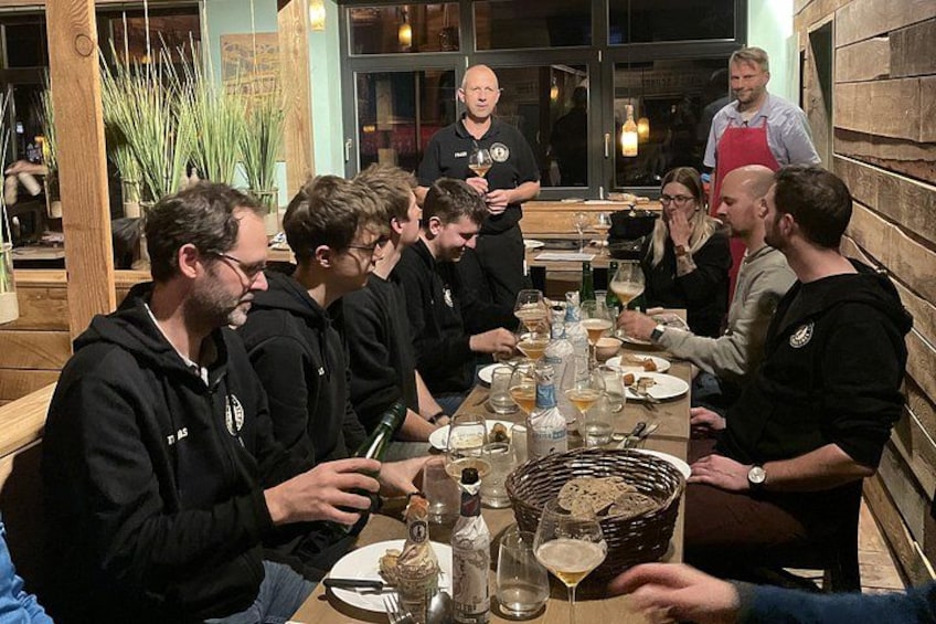 Un grupo de personas sentadas alrededor de una larga mesa en un restaurante con paredes de madera y plantas al fondo. 1