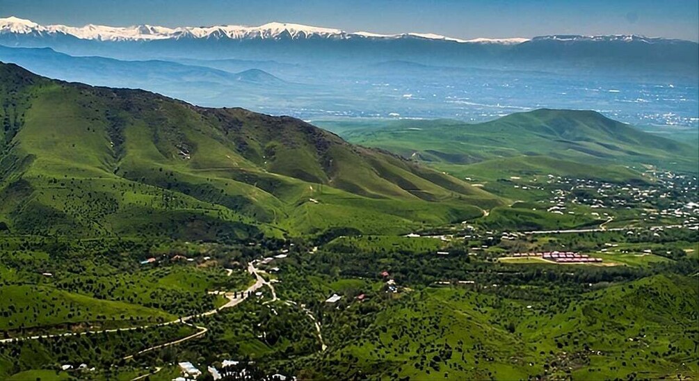 A mountainous landscape with a winding road, scattered buildings, and distant snow-capped peaks. 1