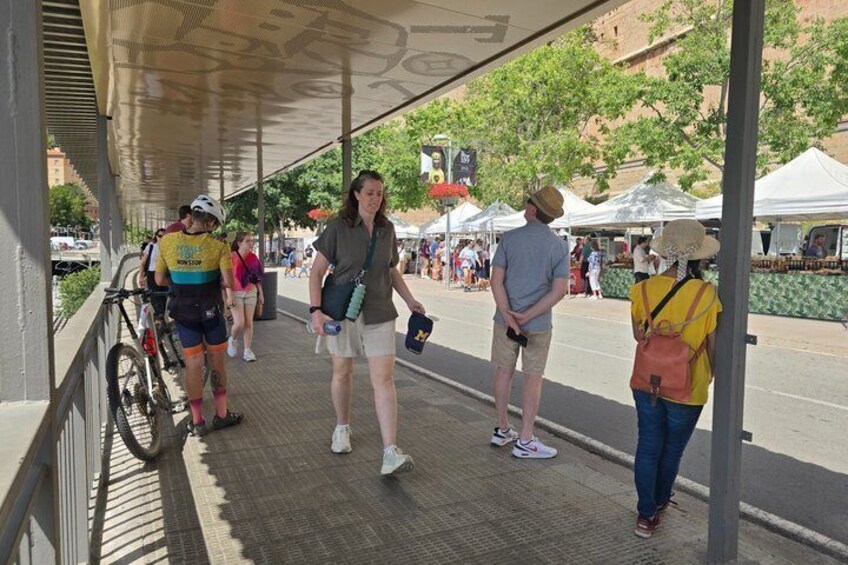 A covered walkway with people standing and walking, a bicycle, and tents in the background. 4