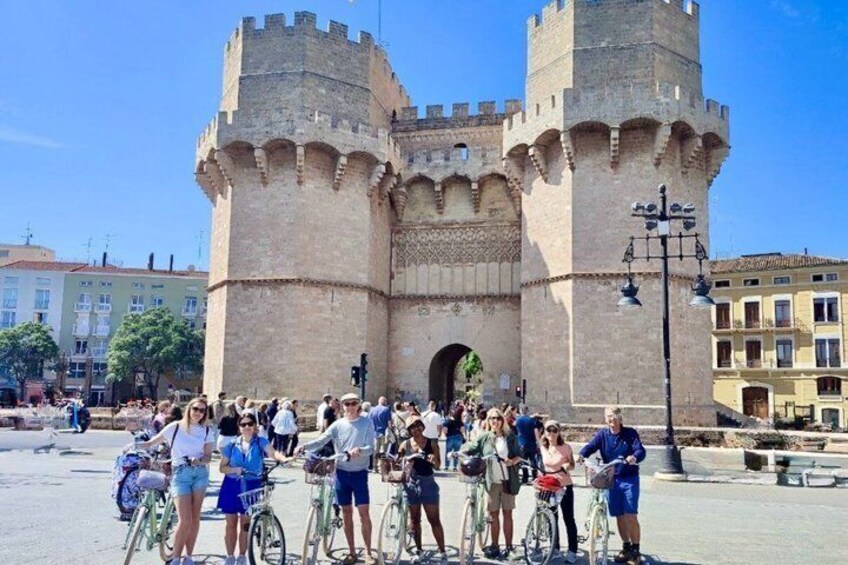En historisk stengate med trappetårn, omgivet af en plads med mennesker og cykler. 1