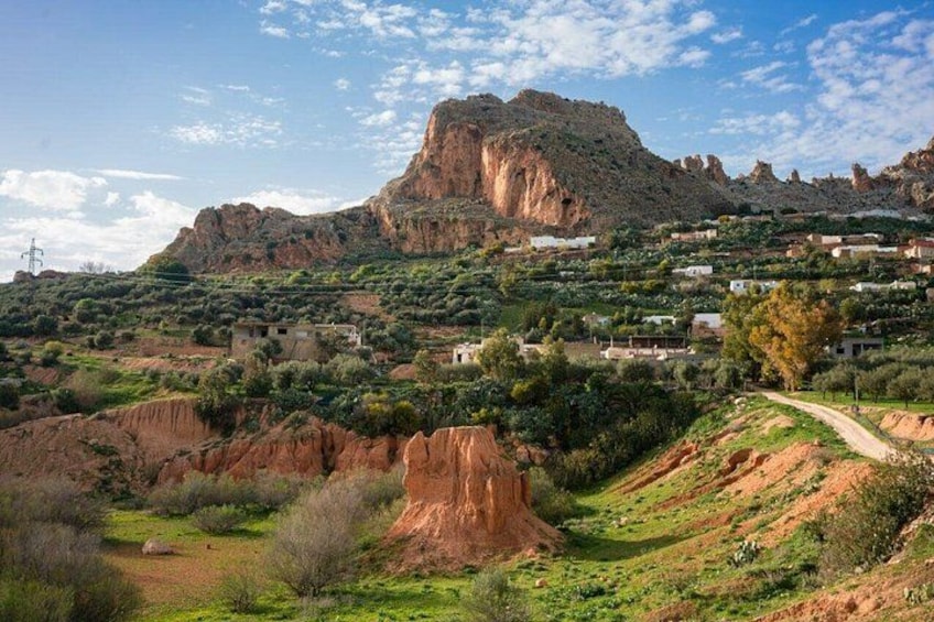 Un paisaje montañoso con una formación rocosa destacada, un camino sinuoso y edificios dispersos. 4