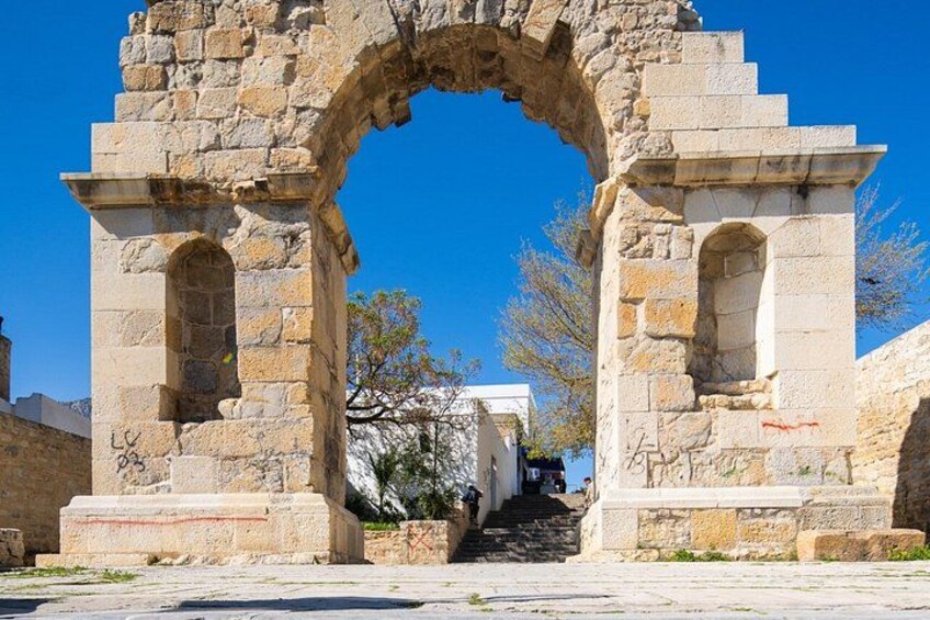 Un antiguo arco de piedra con escaleras que conducen hacia él, rodeado de un cielo azul despejado y algunos árboles. 3