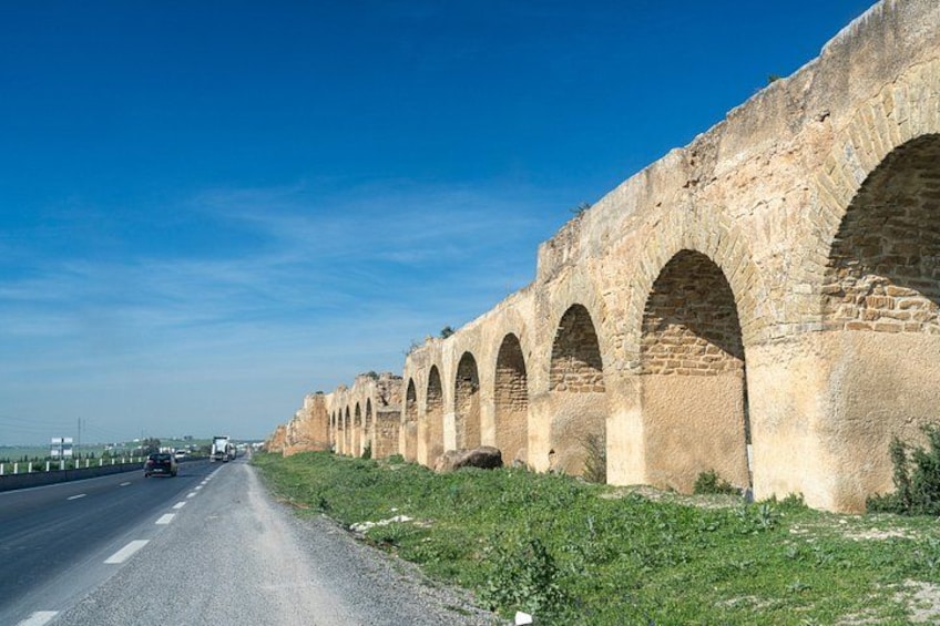 Un largo viaducto de piedra con múltiples arcos, una carretera que lo acompaña y un cielo azul despejado. 1