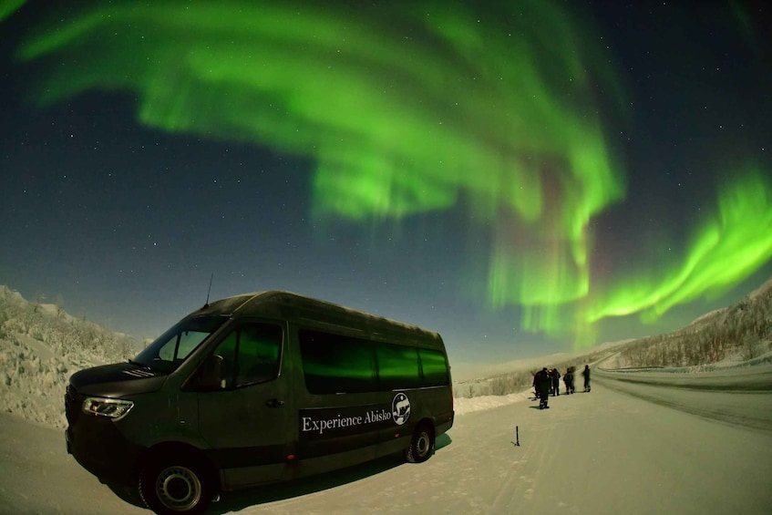A van with 'Experience Abisko' on the side parked on a snowy road with people walking in the background and the Northern Lights in the sky. 1
