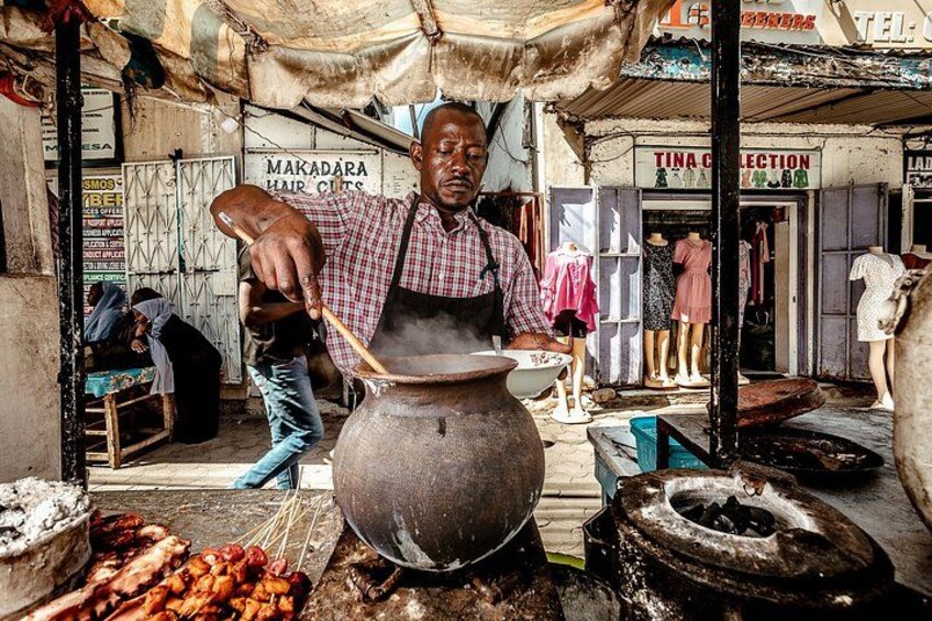 Un vendedor ambulante cocina al aire libre junto a una olla grande, rodeado de varias tiendas y maniquíes. 5