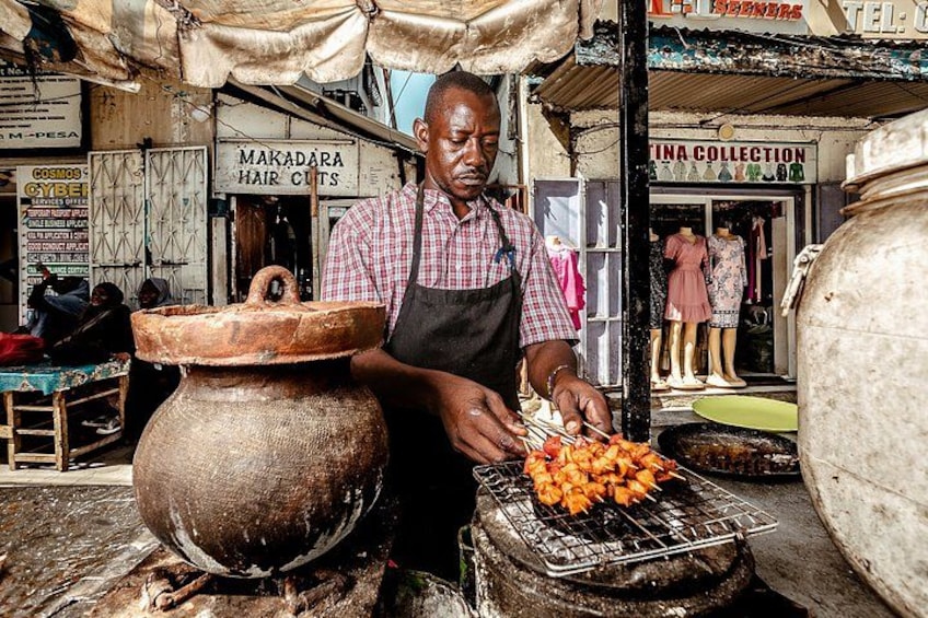 Un vendedor ambulante asando comida al aire libre, con una olla de barro grande y un letrero de corte de pelo al fondo. 1