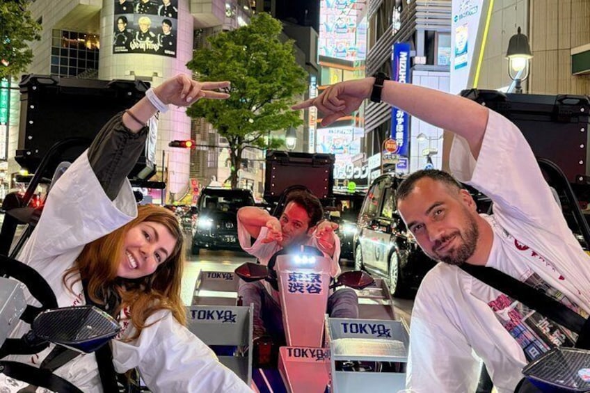 Drei Personen posieren vor einem mit Tokio-Motiven gestalteten Fahrzeug in einer belebten Stadtlandschaft mit Neonreklamen und Werbeanzeigen. 2