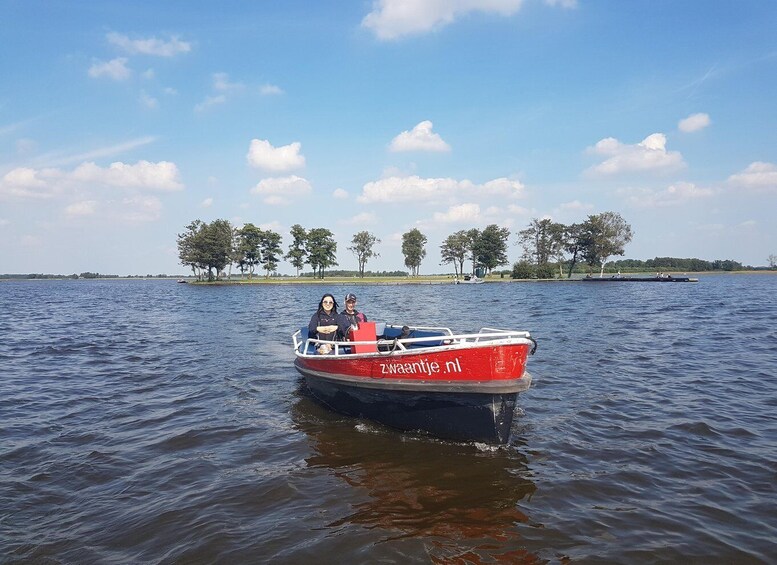 A red boat with 'zwaantje.nl' on the side on calm water, surrounded by trees and a clear sky. 2