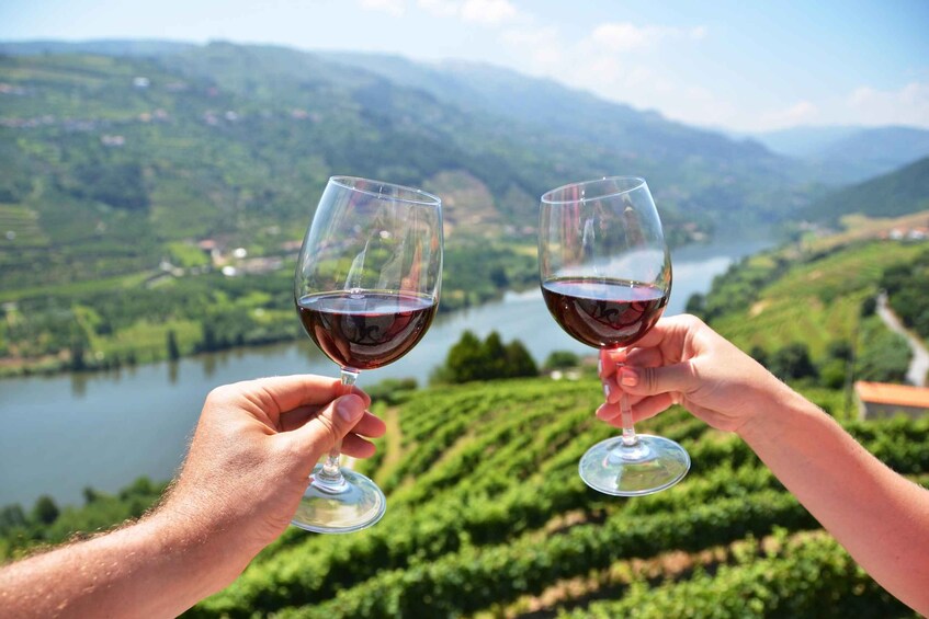 Two hands holding wine glasses with a scenic vineyard and lake in the background. 2