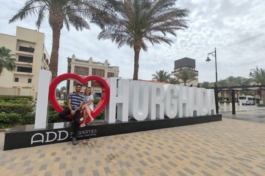 Une enseigne avec le mot « HURGHADA » et une découpe en forme de cœur, des palmiers et des bâtiments en arrière-plan. 1