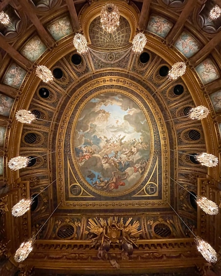 An ornate ceiling with a central fresco, chandeliers, and decorative moldings. 1