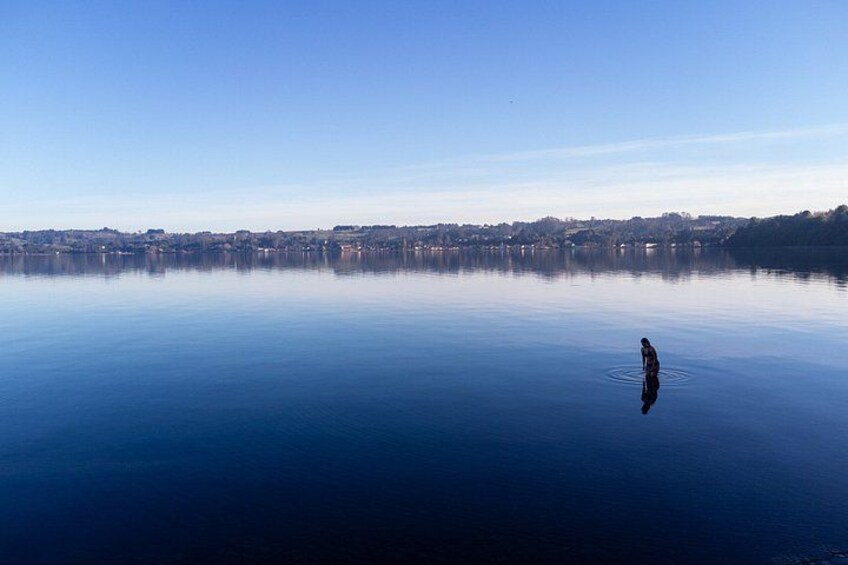 A calm lake with a person standing in the center, surrounded by a clear blue sky and distant buildings. 3