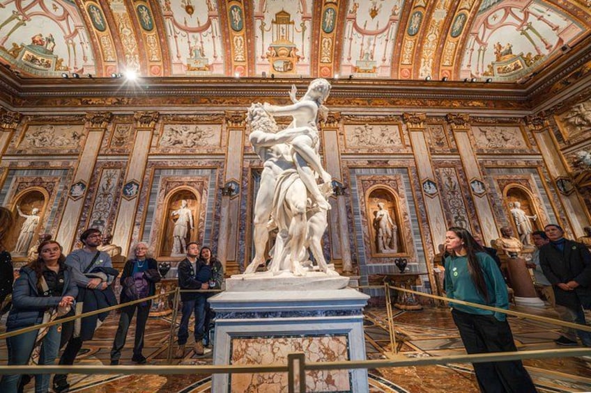 A grand interior with ornate frescoes, a central statue, and visitors observing the artwork. 2