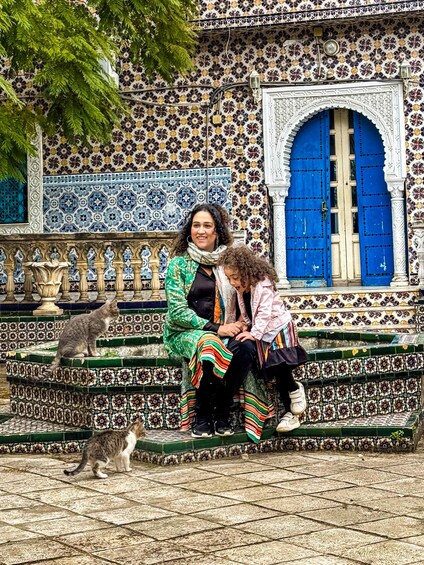 A woman and a child sitting on a tiled bench in front of a building with blue doors and ornate tiles. 4