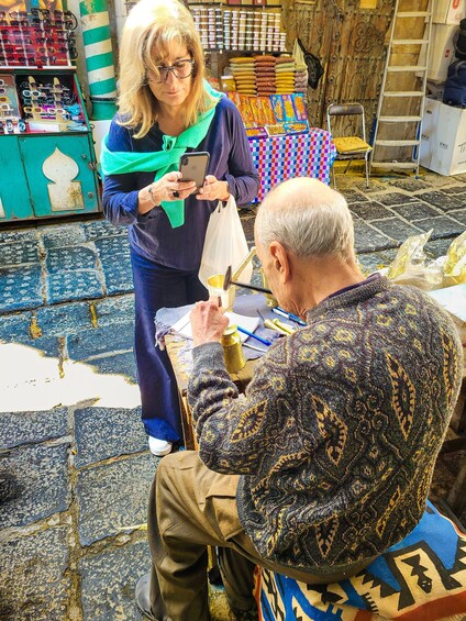 A person sitting at a table with a patterned sweater, another person standing nearby holding a phone, and various items like a ladder and boxes in the background. 5