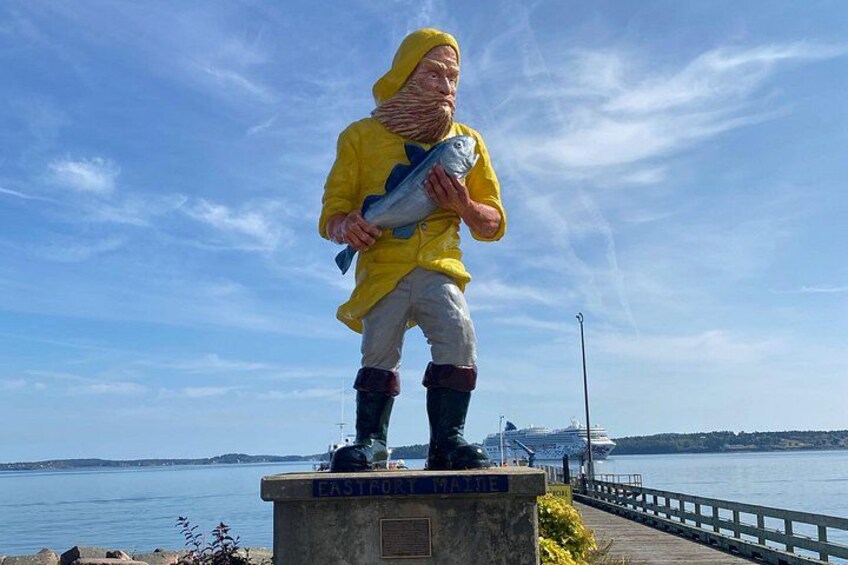 A statue of a person in a yellow raincoat holding a fish, standing on a pedestal with a body of water and a bridge in the background. 2