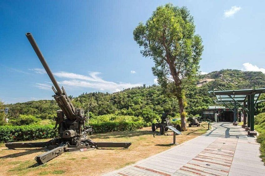 A large artillery piece, a paved walkway, and a covered structure in a park-like setting with trees and hills in the background. 3