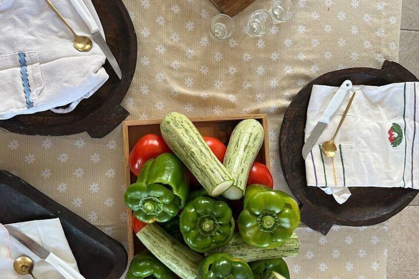 A wooden box filled with green bell peppers, a cucumber, and tomatoes on a patterned tablecloth. 1