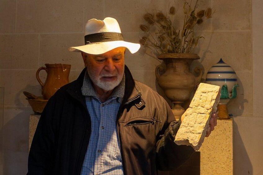 A man holding a stone slab, standing in front of a wall with decorative pottery and a vase. 1