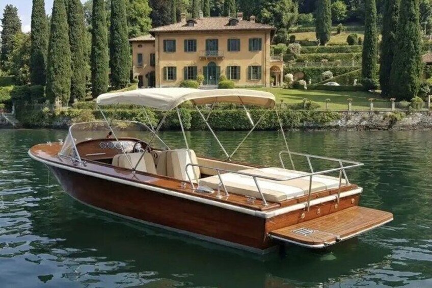 A wooden motorboat with a canopy on a calm lake, surrounded by lush greenery and a large house. 1