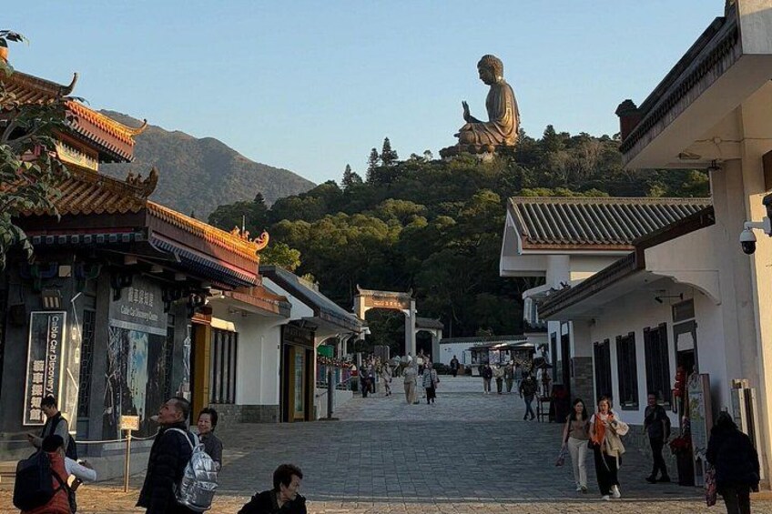 Una calle tradicional con edificios, una gran estatua de Buda en una colina y gente caminando. 3