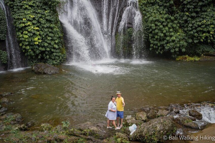 A waterfall with lush greenery and two people standing by the water. 2