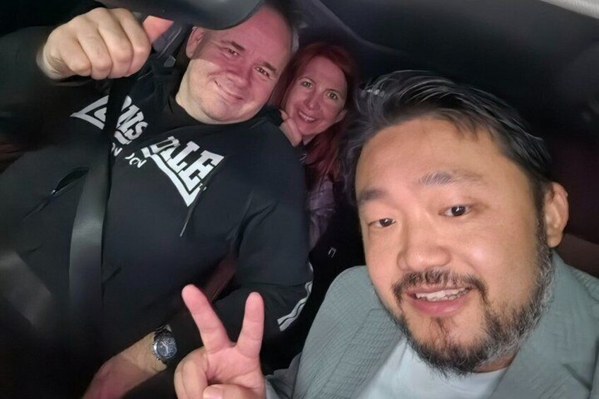 Zwei Personen im Auto, eine trägt ein schwarzes Kapuzenshirt mit weißer Aufschrift, die andere ein helles Hemd. 2