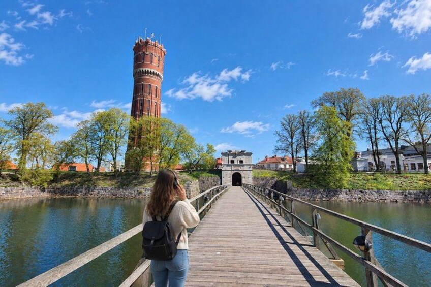 A wooden bridge leading to a historic tower by a calm river, surrounded by trees and buildings. 3