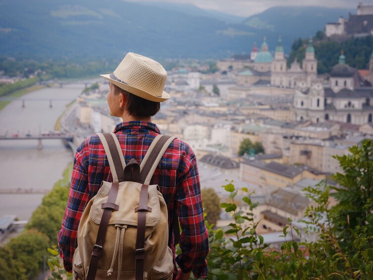 Eine Person mit Rucksack und Hut blickt auf einen Fluss und historische Gebäude. 4