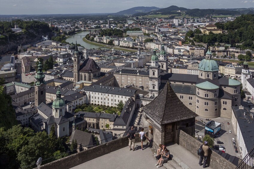 Ein weiter Blick auf eine historische Stadt mit markanter Architektur, darunter ein schlösserähnliches Gebäude mit grüner Kuppel, ein Fluss, der durch die Stadt fließt, und ein Hügel mit einer Kirche auf dem Gipfel. 3