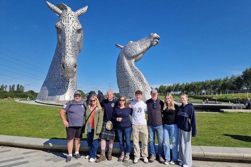 En grupp människor står framför Kelpies, två stora hästskulpturer, med en klarblå himmel i bakgrunden. 1
