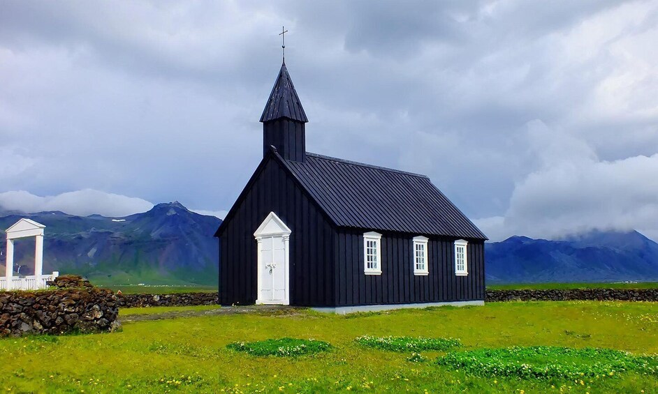 A black church with a white door and windows, a steeple, and a stone wall in a grassy field with mountains in the background. 2