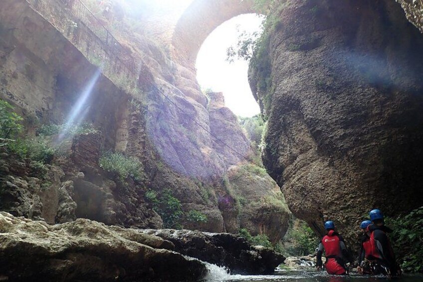 A canyon with a narrow water stream, rocky terrain, and two people wearing helmets and red jackets. 4