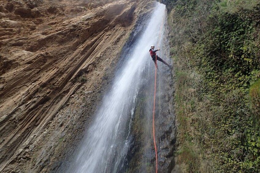A person rappelling down a steep, rocky cliff with a waterfall in the background. 1