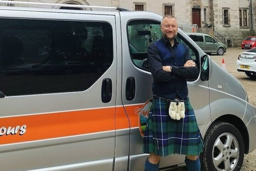 A man in a kilt standing next to a silver van with orange stripes and the word 'COTTON' on it. 1