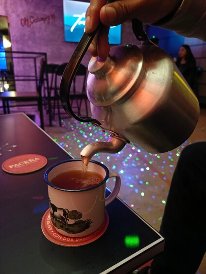 A hand pouring tea from a metal teapot into a mug with a rooster design. 2