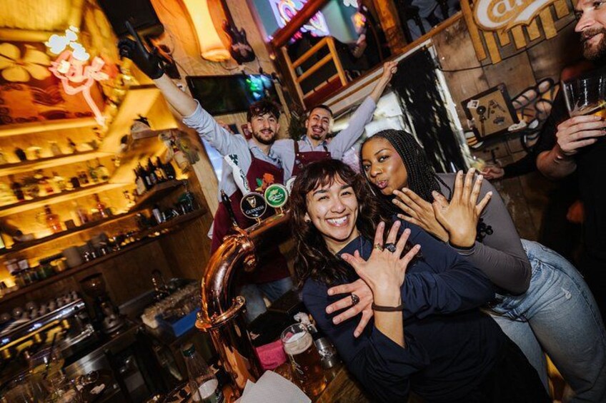 A bar with shelves of bottles, a large copper beer tap, and people celebrating. 2