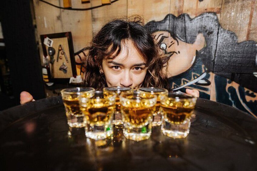A person holding a tray with multiple shot glasses filled with a golden liquid, likely alcohol, in a dimly lit indoor setting with graffiti on the wall. 2