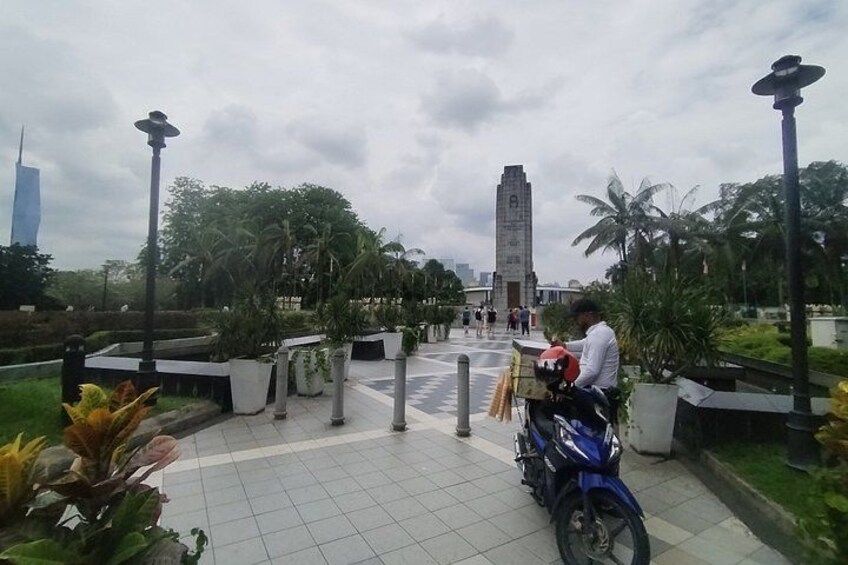 Un homme est assis sur une moto bleue dans un parc, avec un grand monument en arrière-plan. 2